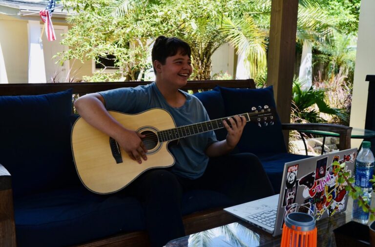 A student plays an acoustic guitar on a blue couch on the covered patio